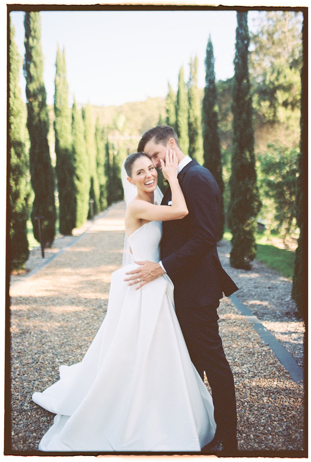 Newlyweds share an intimate moment on a tree-lined path with cypress trees in warm sunlight.