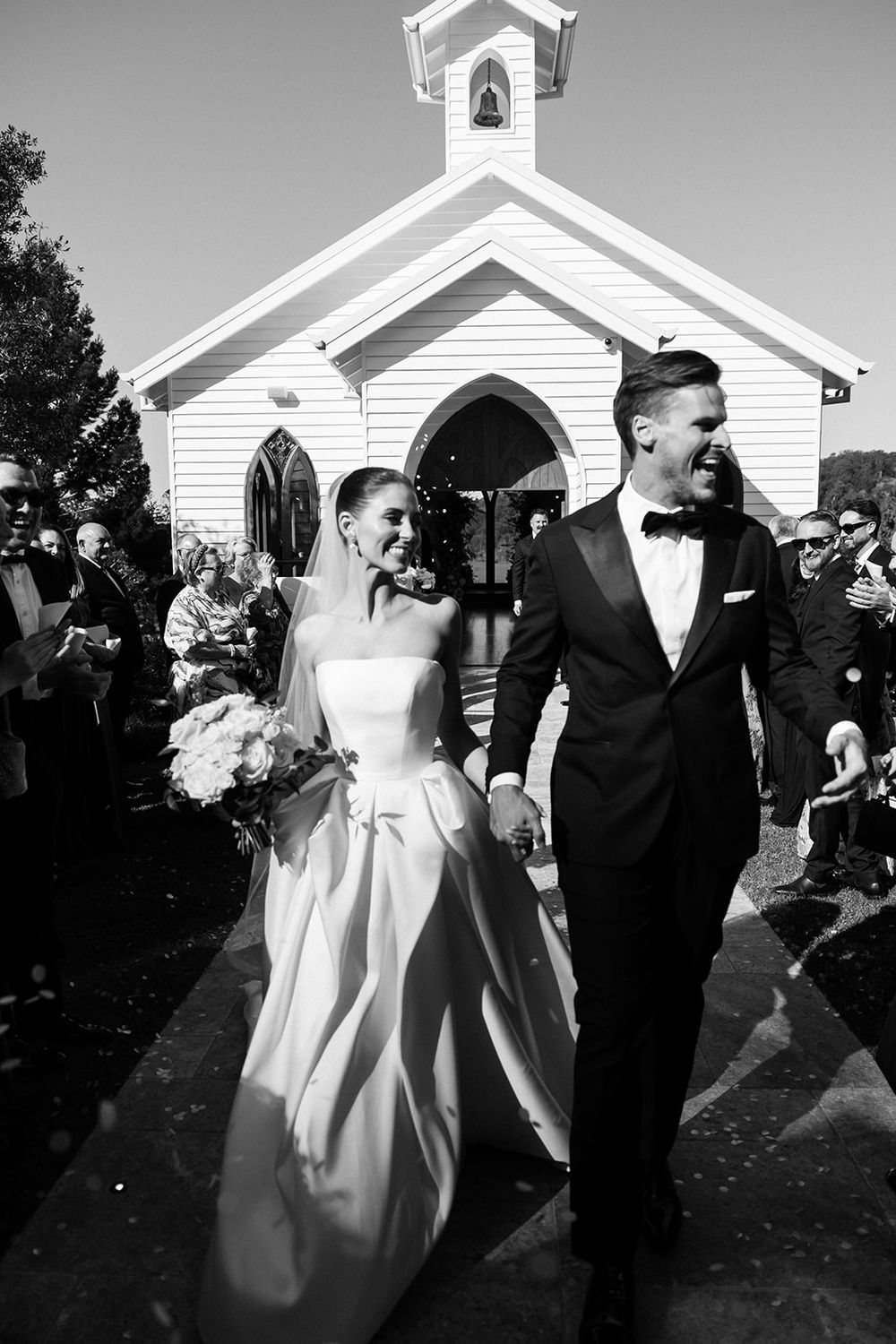 A black and white wedding photo of newlyweds exiting a small white chapel surrounded by guests.