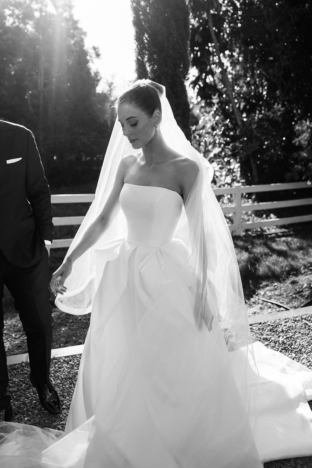 Elegant strapless white wedding gown with flowing veil captured in black and white photography during an outdoor ceremony.