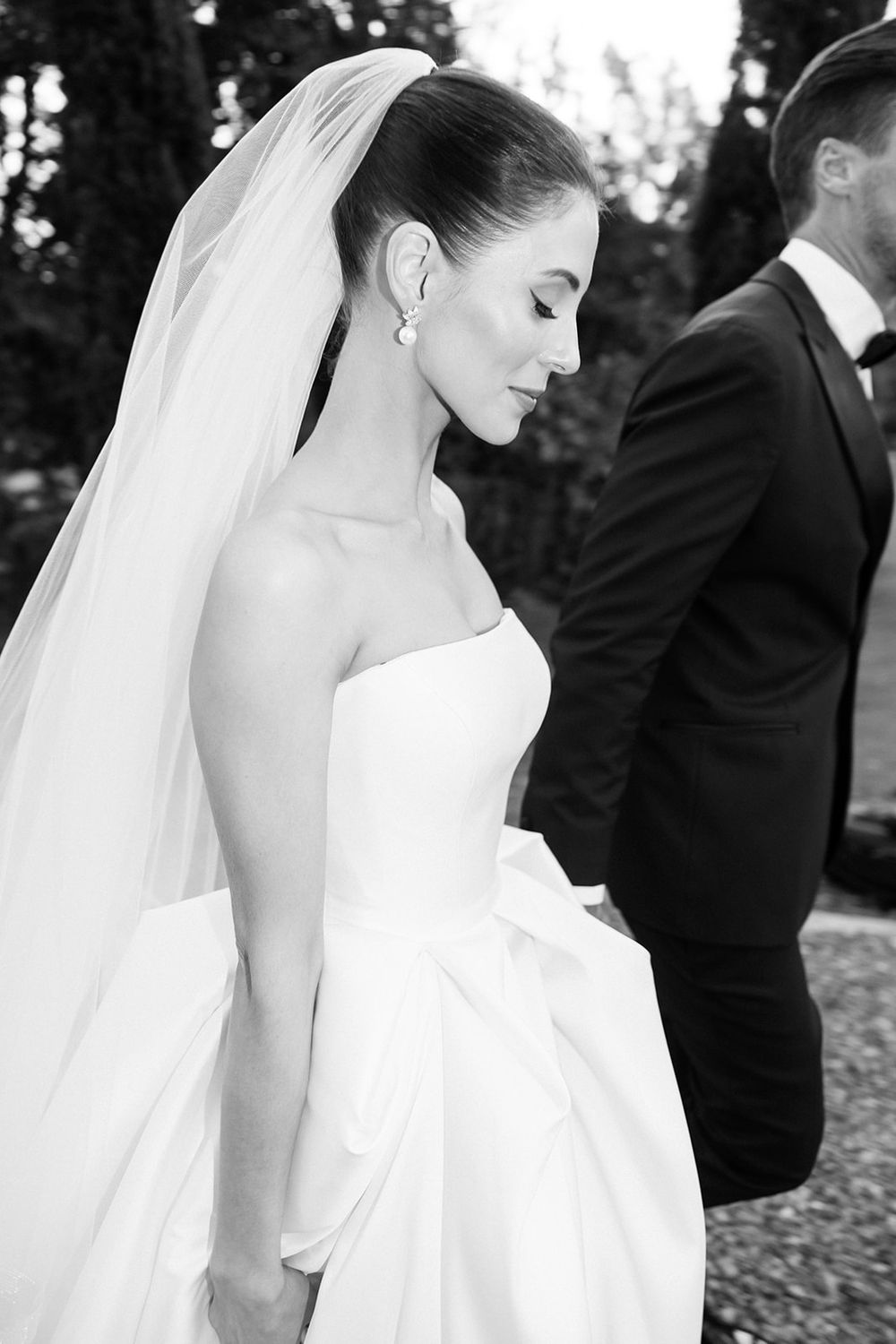 Black and white portrait of a bride in a strapless wedding gown with flowing veil as groom stands in background.