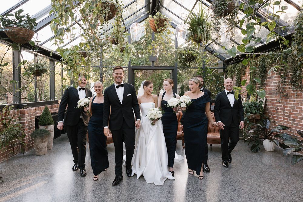 A wedding party celebrates with joyful jumping poses inside a beautiful glass conservatory during a formal ceremony.