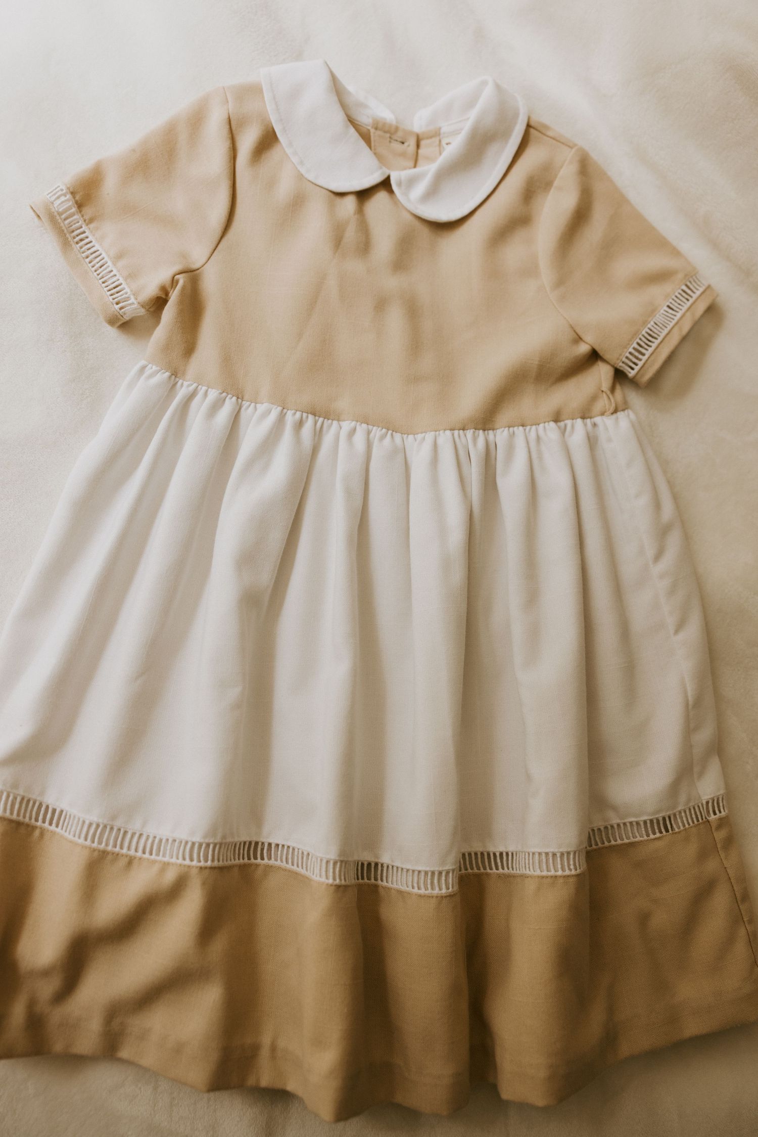 Formal infant outfit with beige vest, white shirt, light blue bow tie, and matching shorts hanging on wooden ladder shelf.