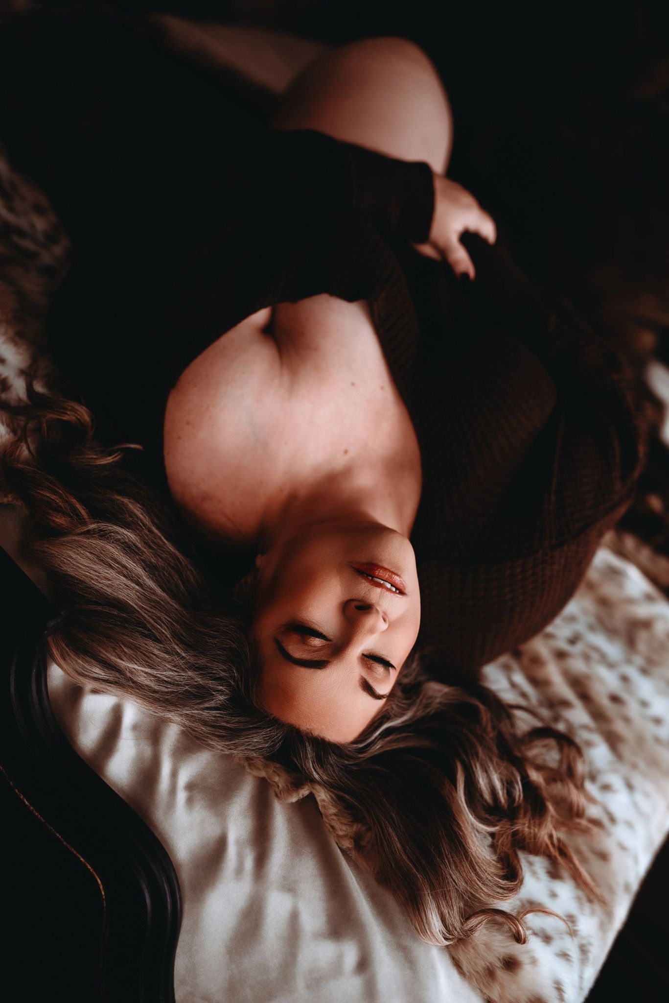 Artistic boudoir photography in dark tones shows person with long wavy hair lying on white bedding.