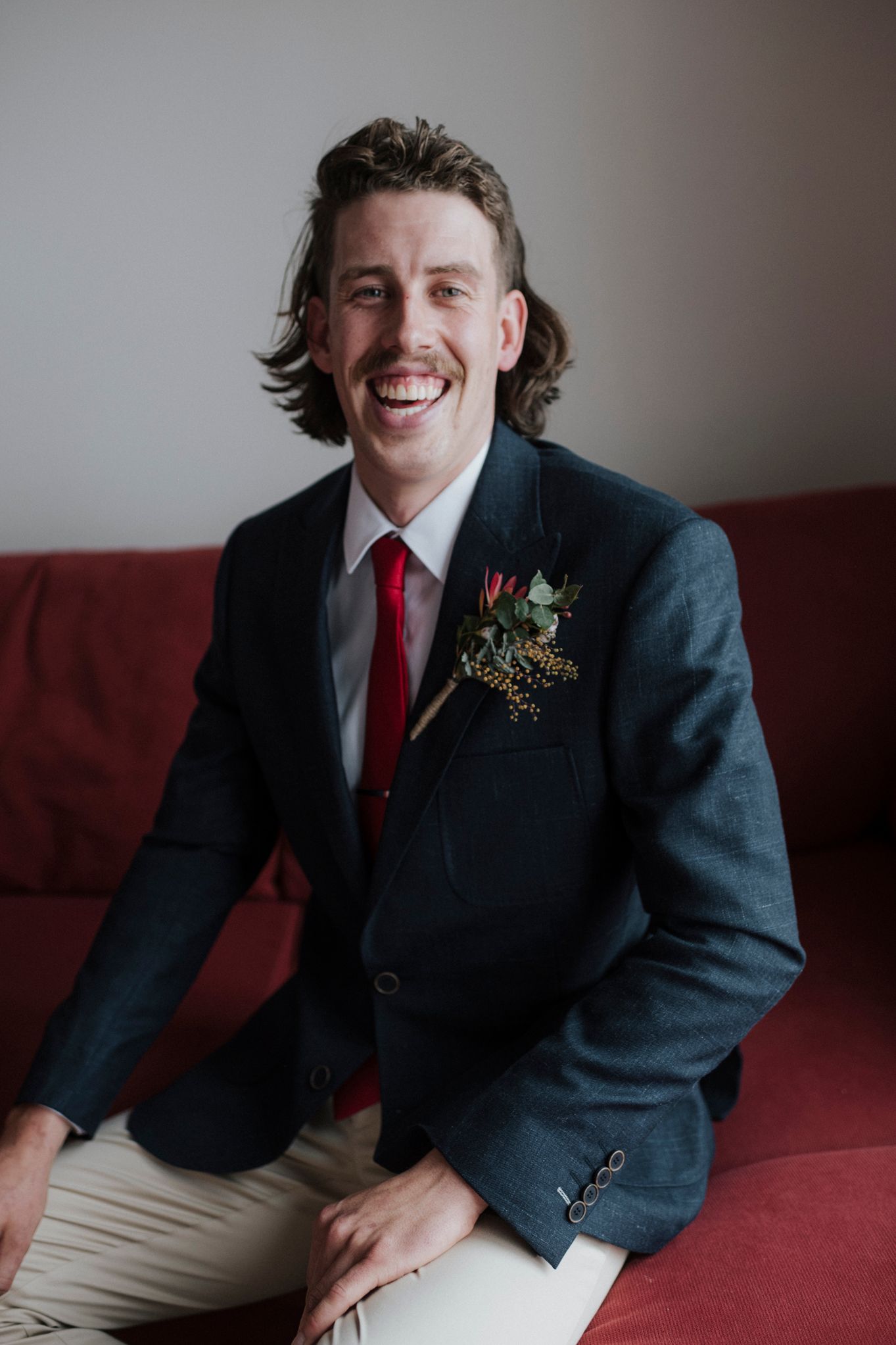 A groom in a navy blazer and red tie smiles while seated on a red couch with a floral boutonniere on his lapel.