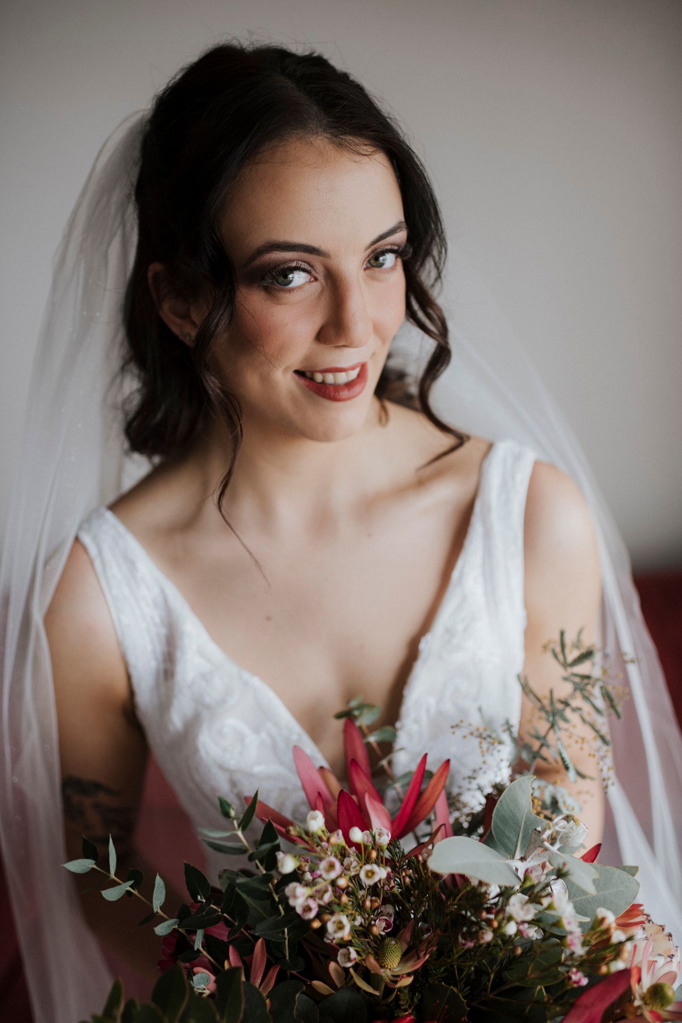 Bride in white dress holds vibrant red and green bouquet while wearing a delicate veil against soft lighting.