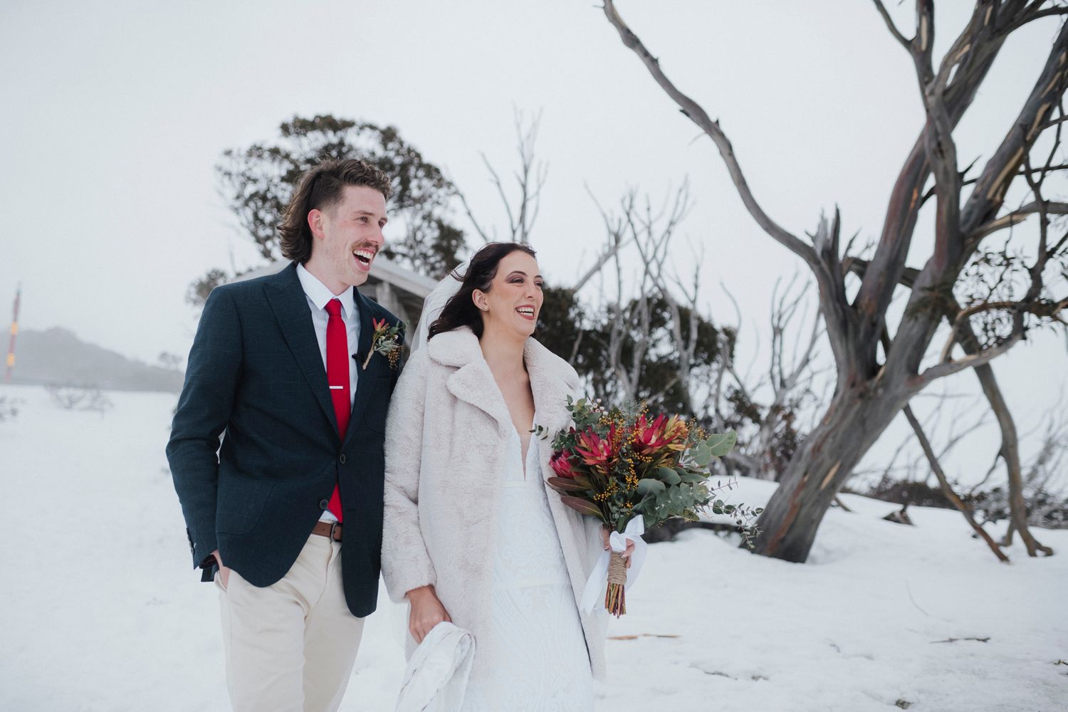 A couple celebrates their winter wedding in a snowy mountain landscape with stark trees in the background.