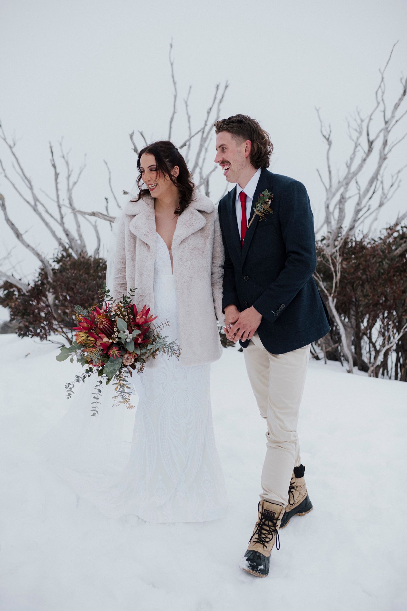Couple shares candid moment together in a snowy winter landscape with red floral bouquet.