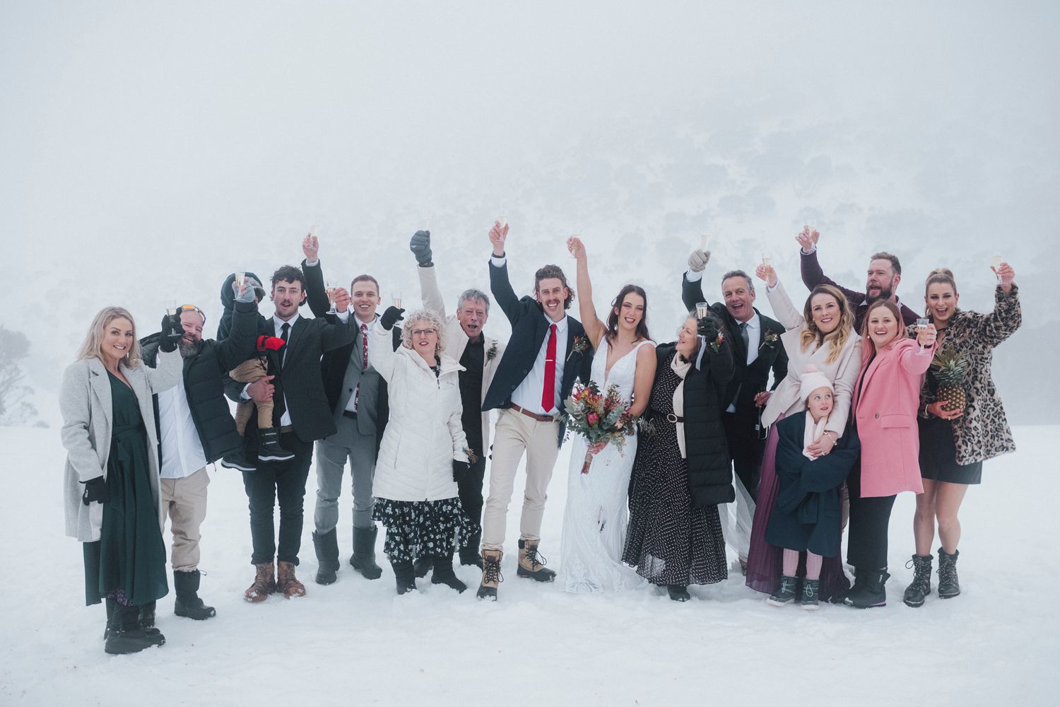 A wedding party dressed in formal attire poses together in a snowy winter landscape.