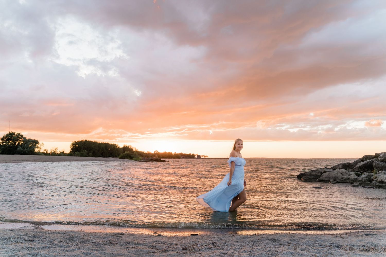 Springfield High School senior in a flowing white dress stands on a rocky beach at sunset with orange clouds reflecting on the water.