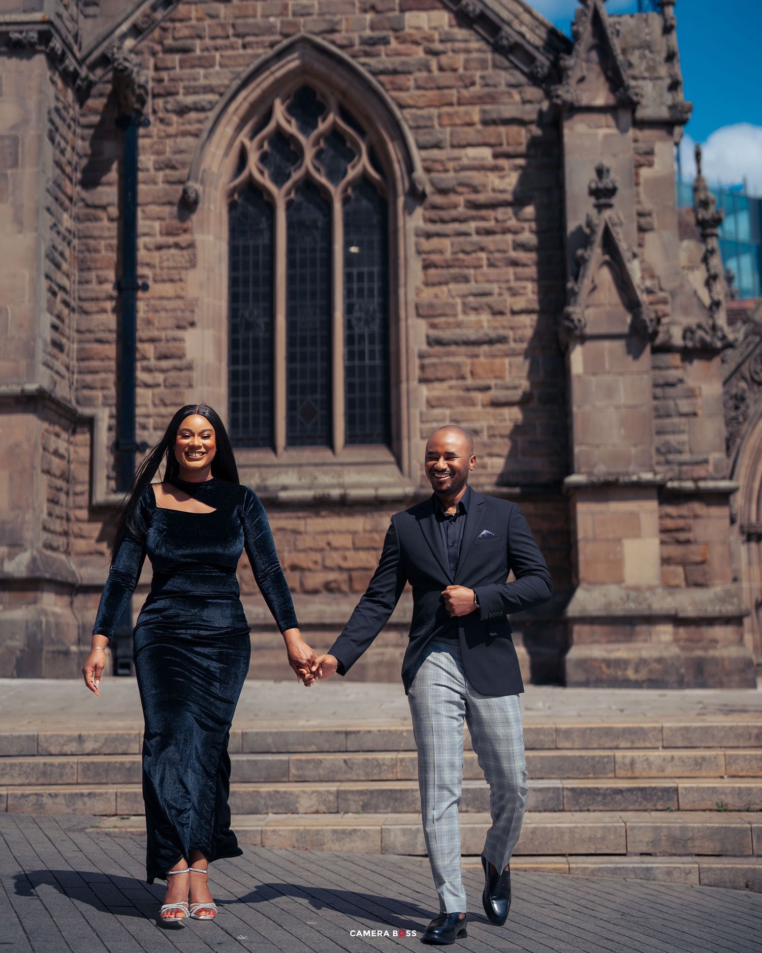A couple holds hands while walking down stone steps in front of a Gothic church with arched windows on a sunny day.