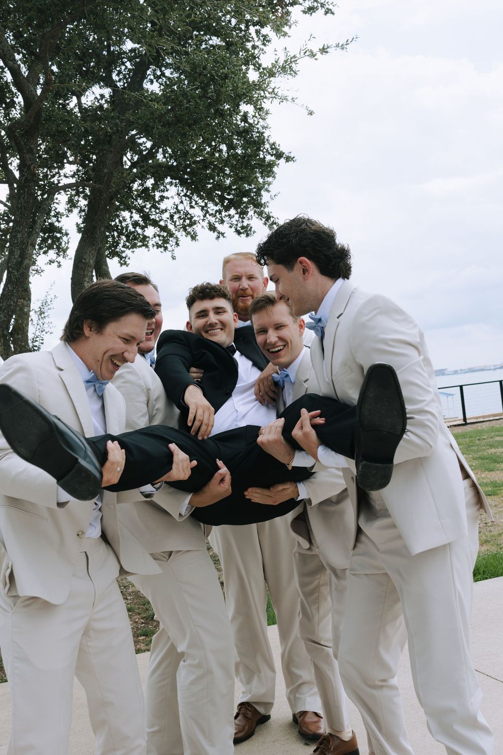 Groomsmen in light colored suits laughing together in a candid line-up photo outdoors.