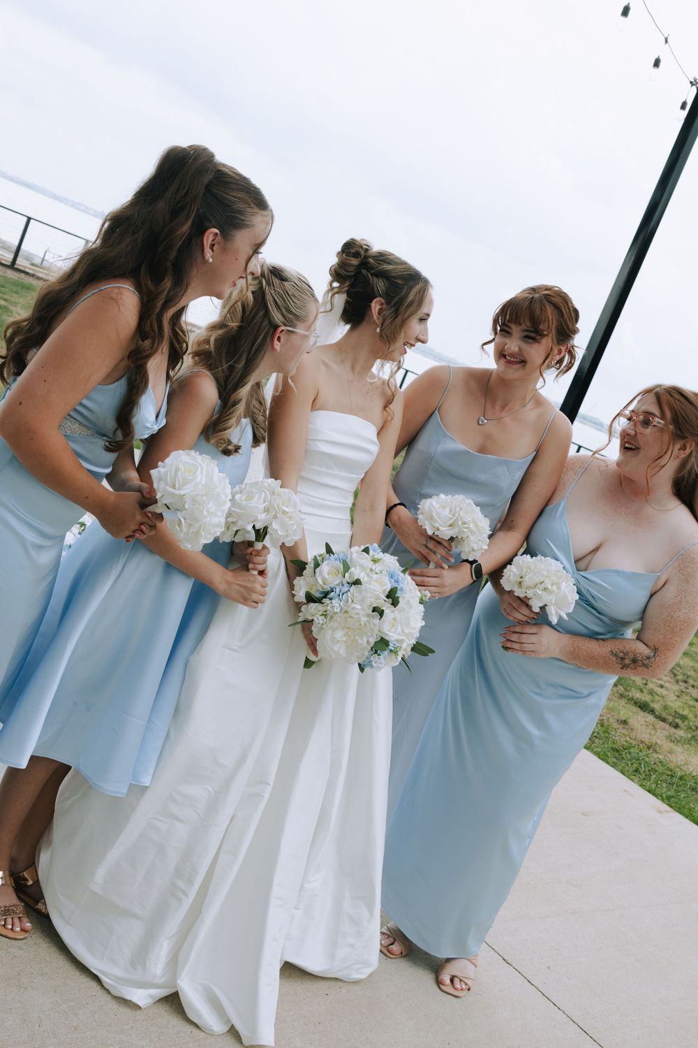 Bridesmaids in light blue dresses and bride in white gown standing together outdoors.