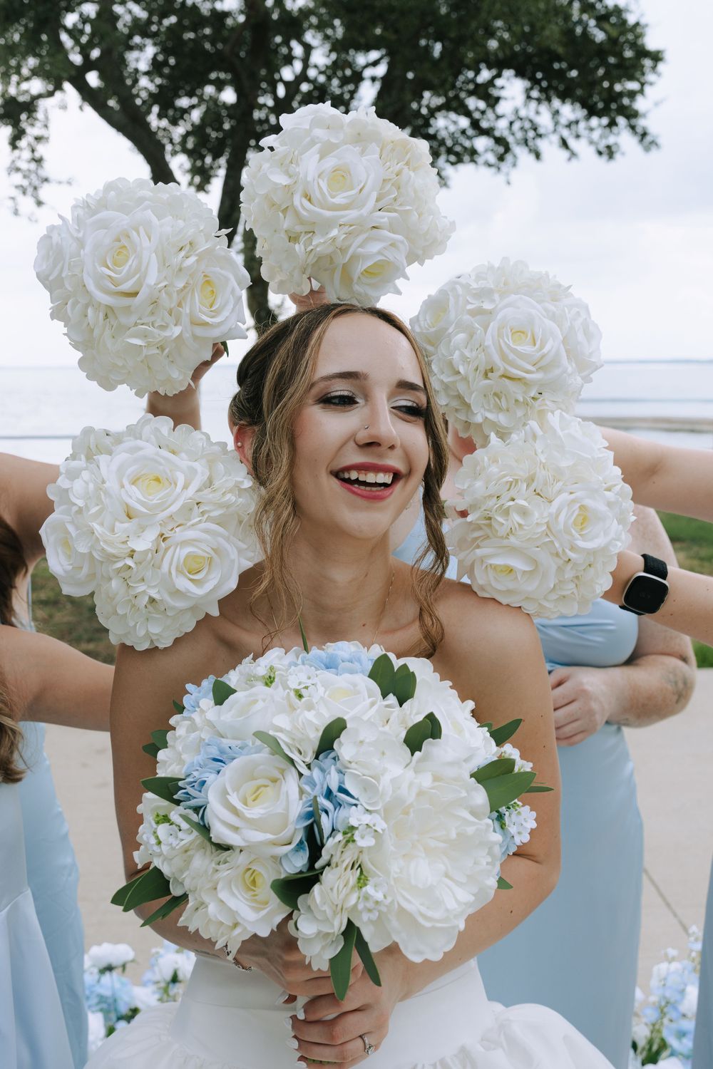 A joyful bride in a white dress holds her bouquet while bridesmaids hold white rose pomanders behind her at an outdoor wedding.