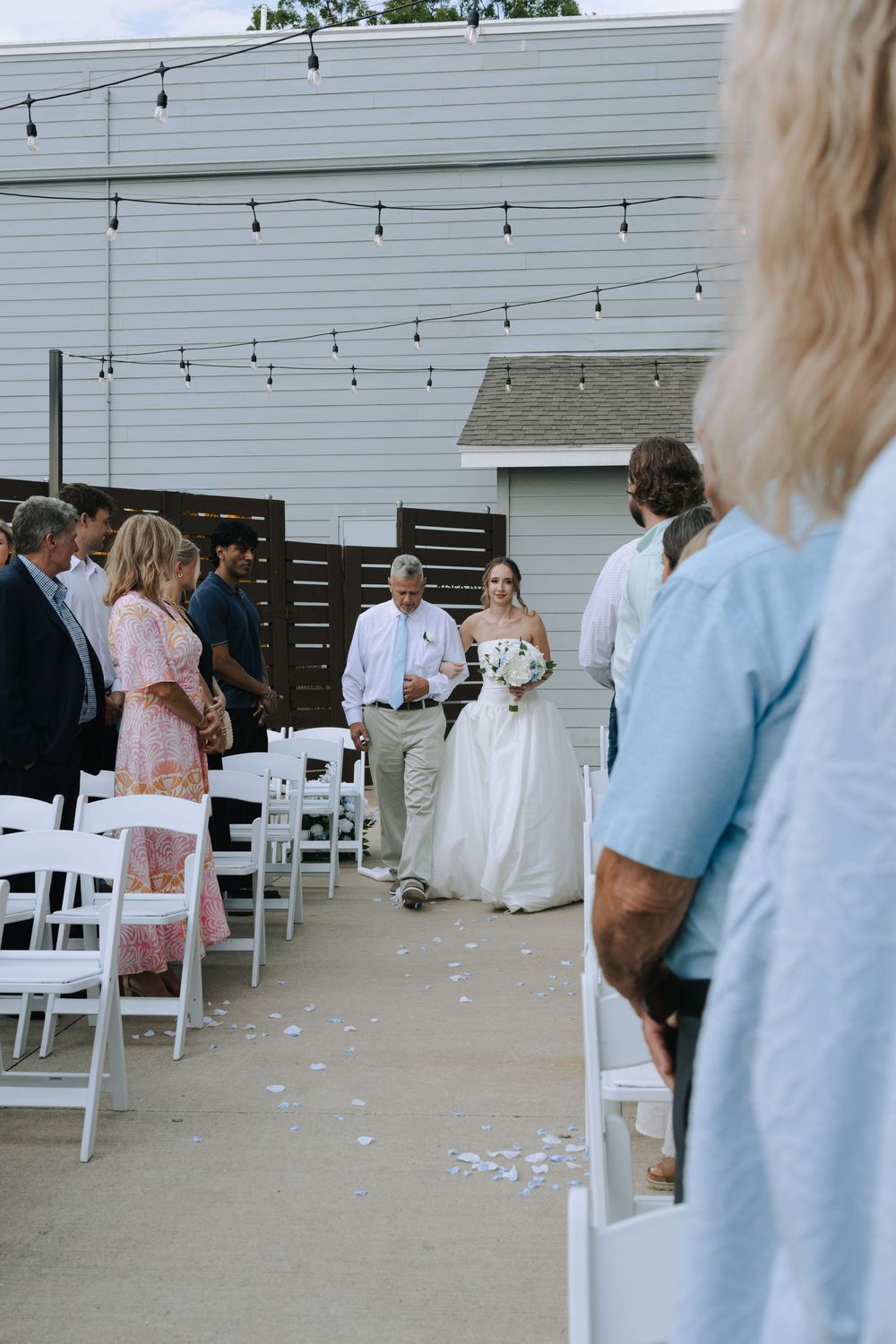 Outdoor wedding ceremony setup with white chairs and hanging string lights against a white building.