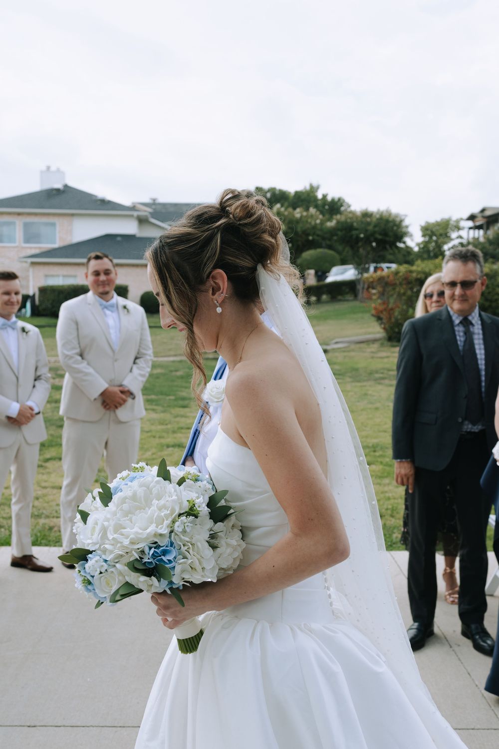 A bride in a white wedding dress holds a bouquet of white flowers while standing outside with guests in formal attire.