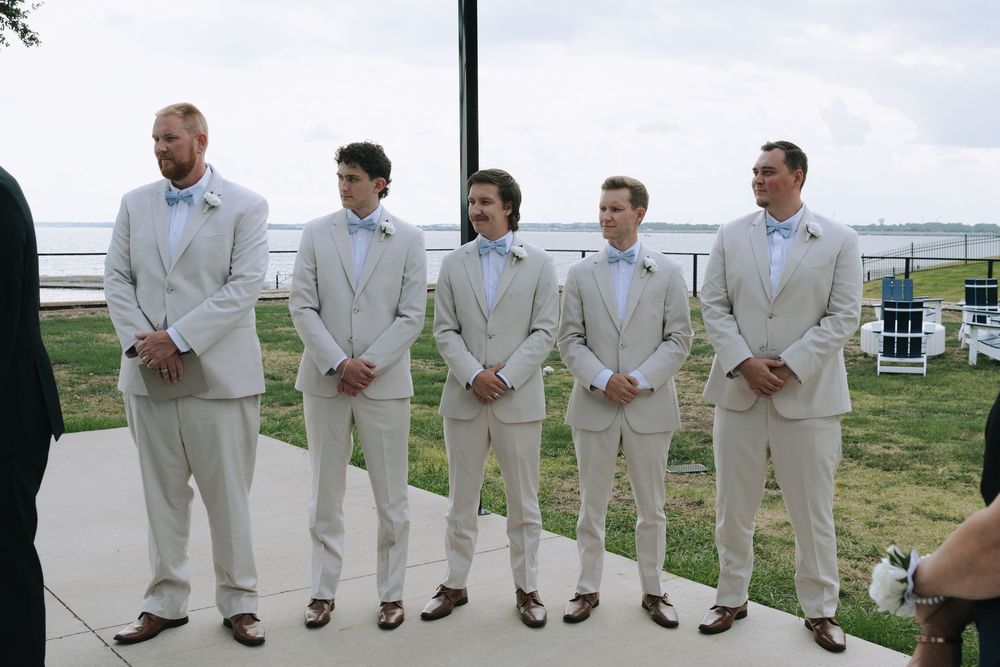 Wedding party in crisp white suits lined up outdoors by the water.