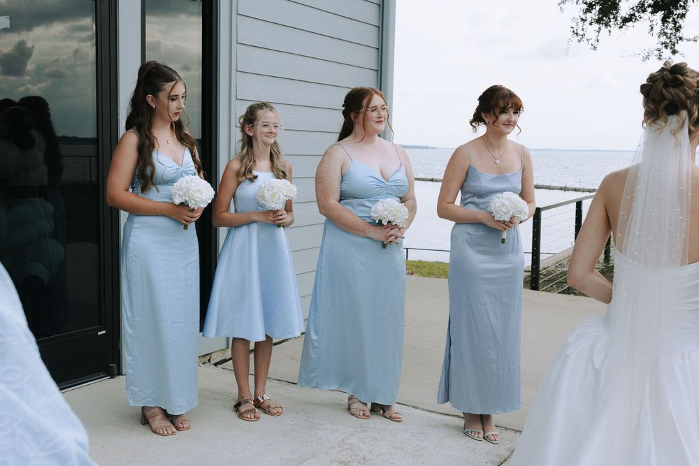 Bridesmaids in light blue dresses stand in a row on a white porch during a wedding.
