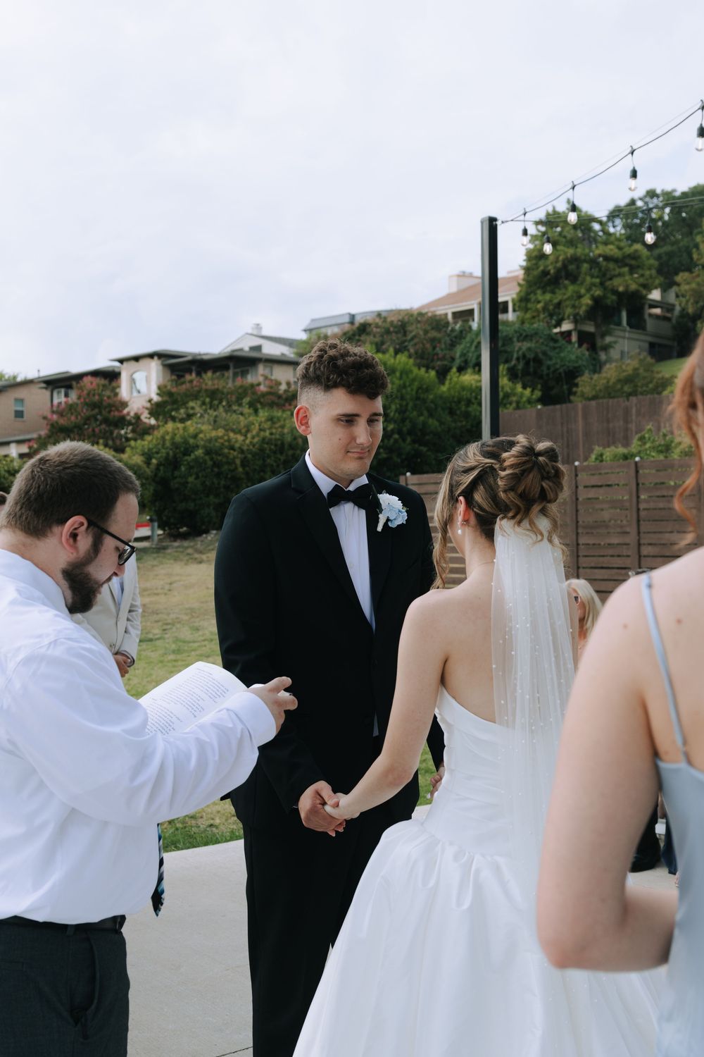 Outdoor wedding ceremony taking place on a paved area with string lights and hillside homes in the background.
