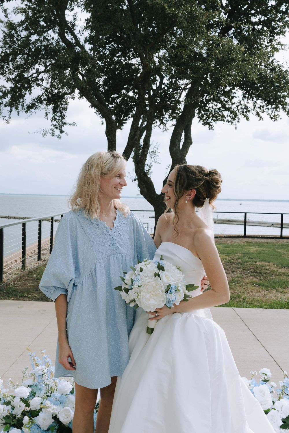 Two people share a tender moment outdoors near the ocean, one wearing a white dress and the other in light blue.