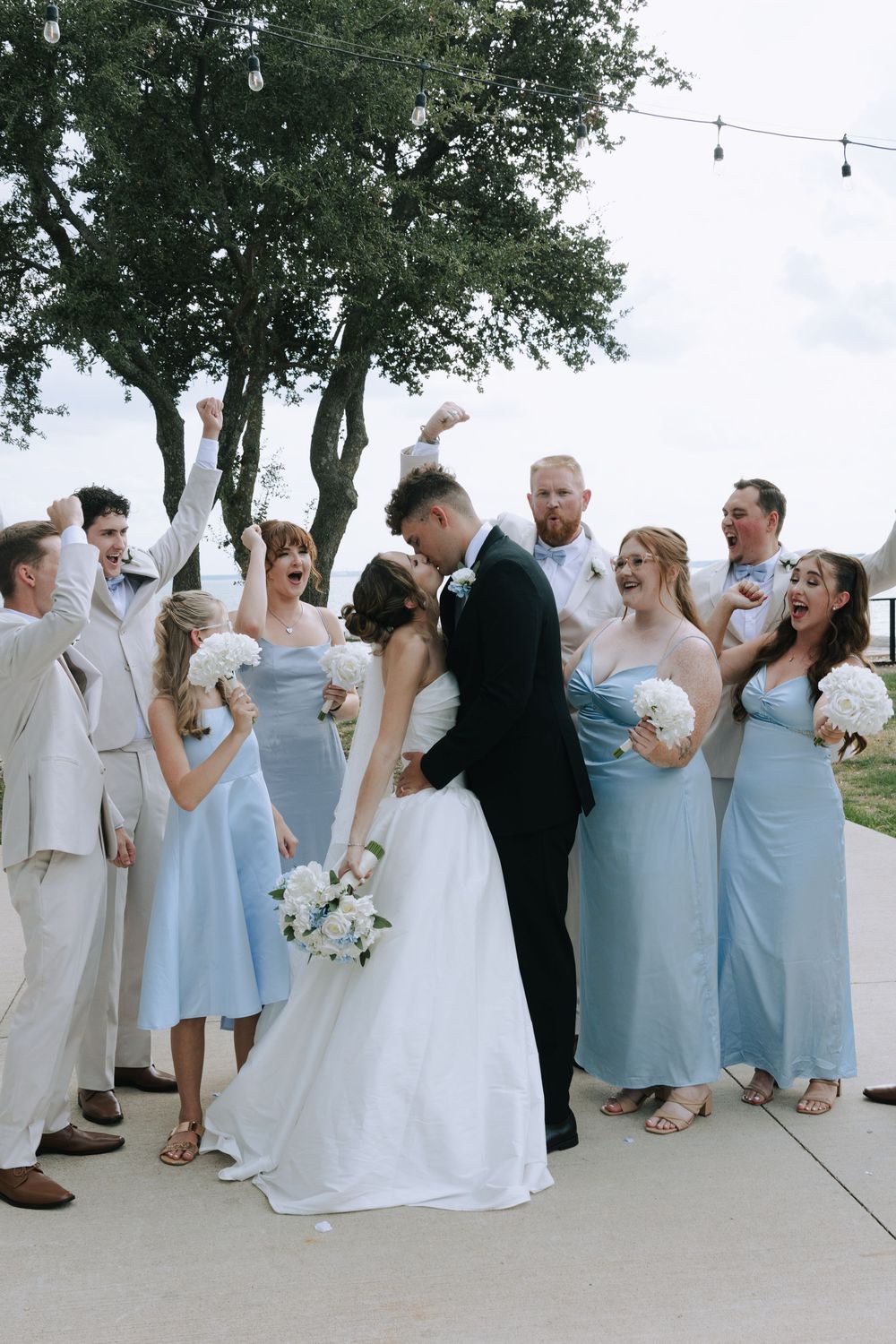 Wedding party in light blue dresses and cream suits celebrates outdoors under string lights.