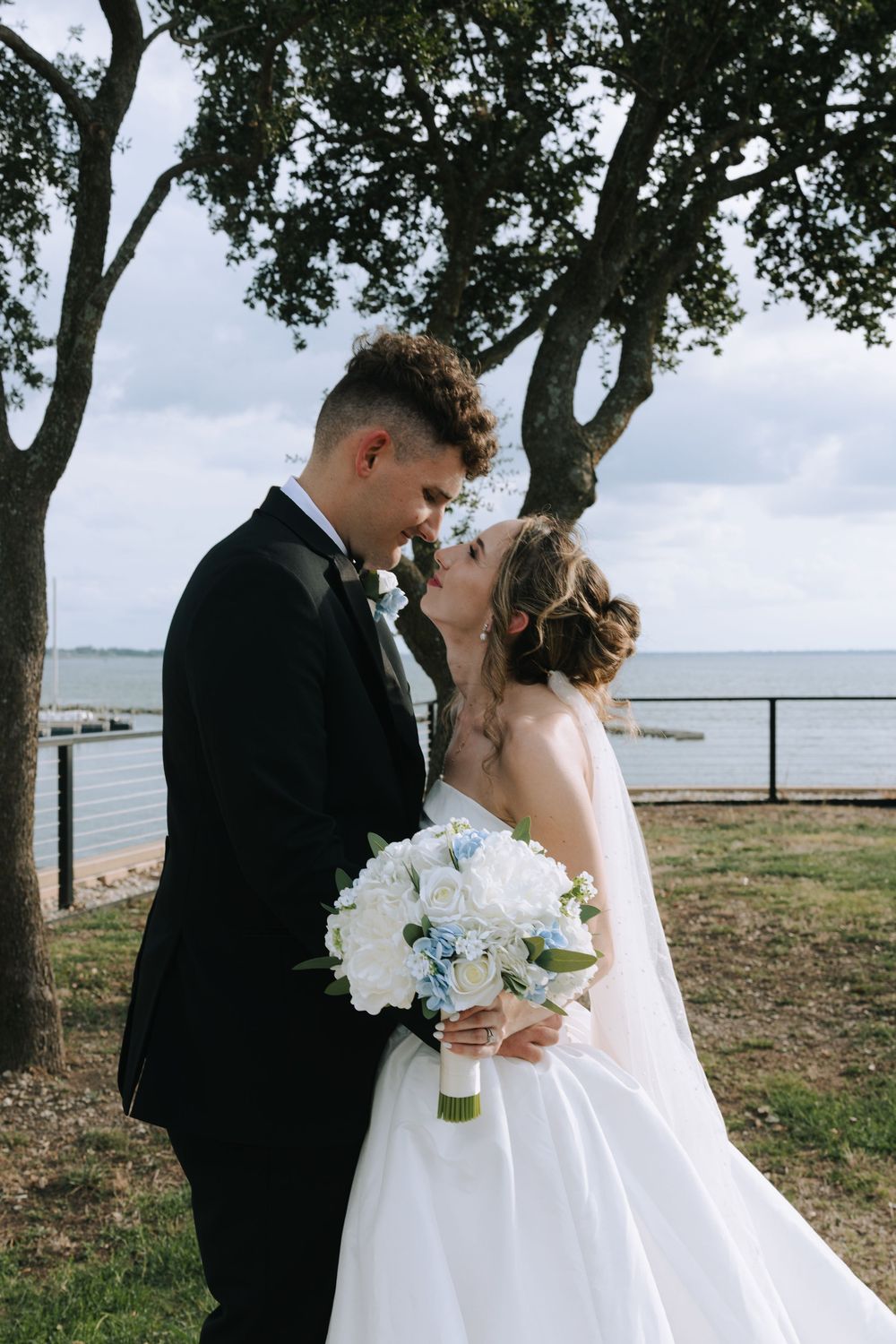 A romantic wedding moment captured under trees by the water with the couple sharing a tender kiss.