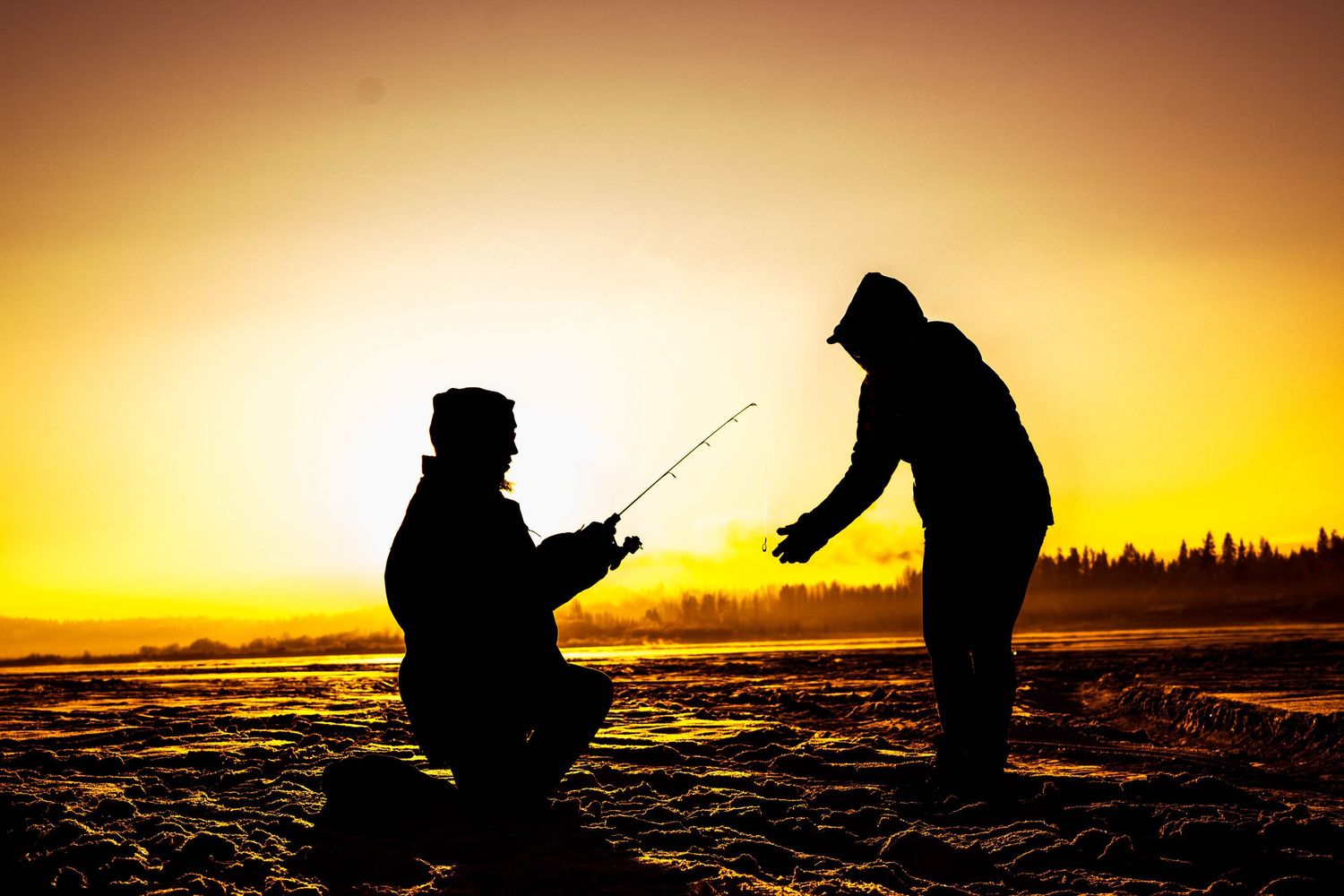Two silhouetted figures fishing together on a beach at sunset with dramatic golden sky backdrop.