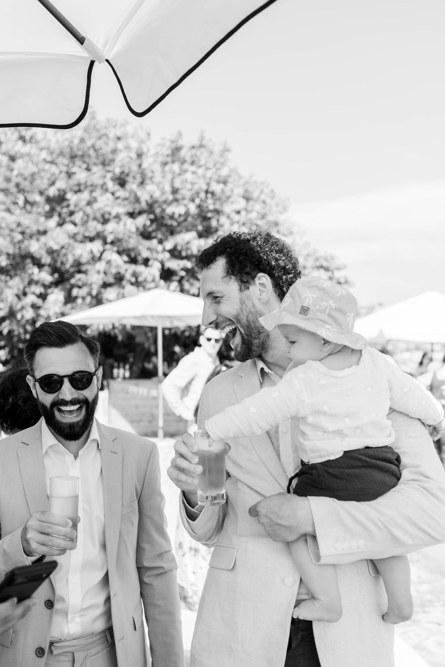 Schwarz-Weiß-Foto von zwei Männern und einem Baby bei einer Hochzeit auf Mallorca, die gemeinsam lachen, weil das Kind die Hand im Bier hat.