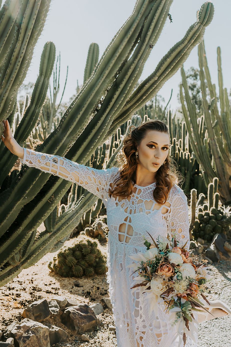 Ethereal bride in lace gown poses dramatically against towering saguaro cacti in desert landscape with floral bouquet.