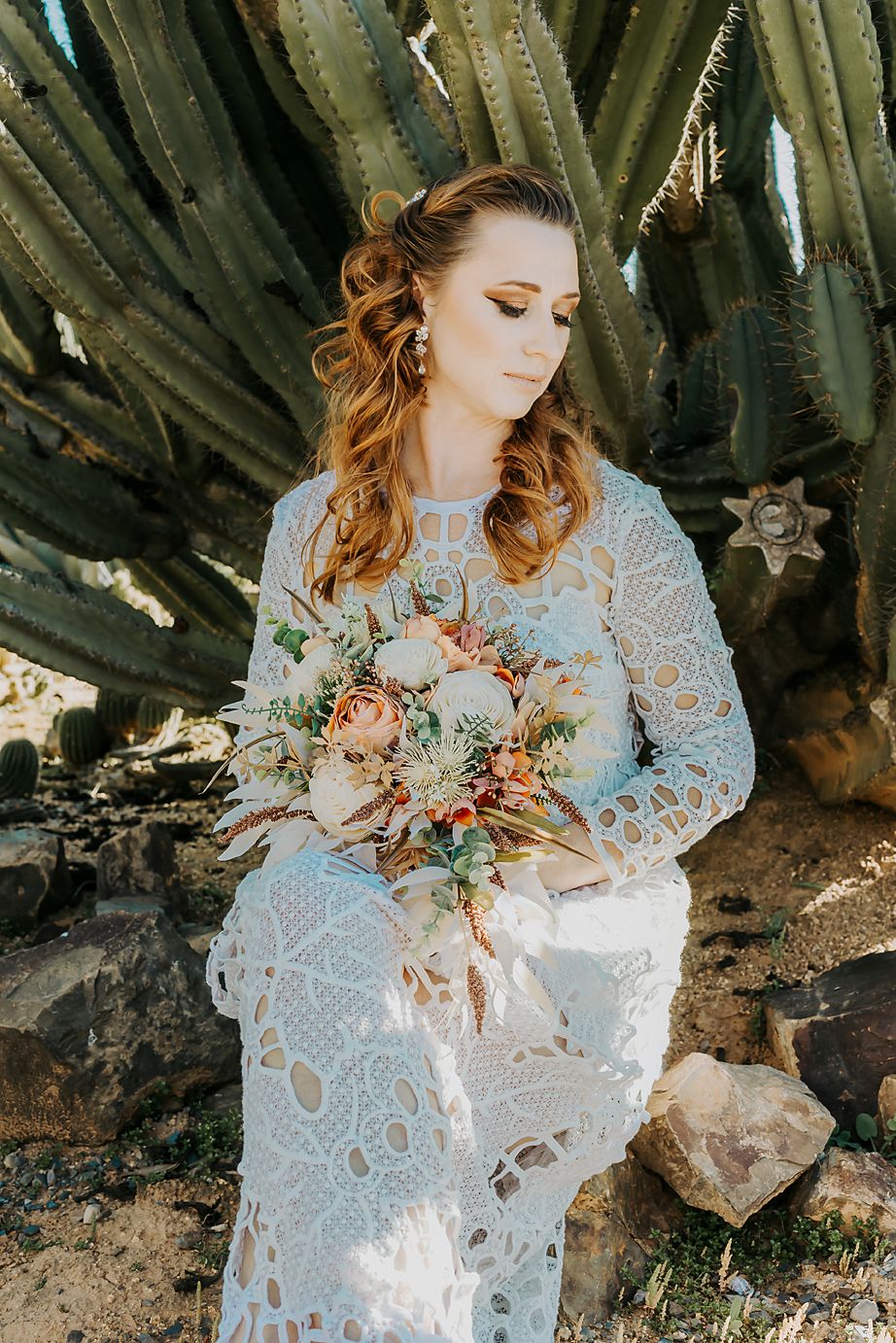 A bride in an elegant lace gown poses gracefully against desert cacti while holding a romantic bouquet of pink flowers.
