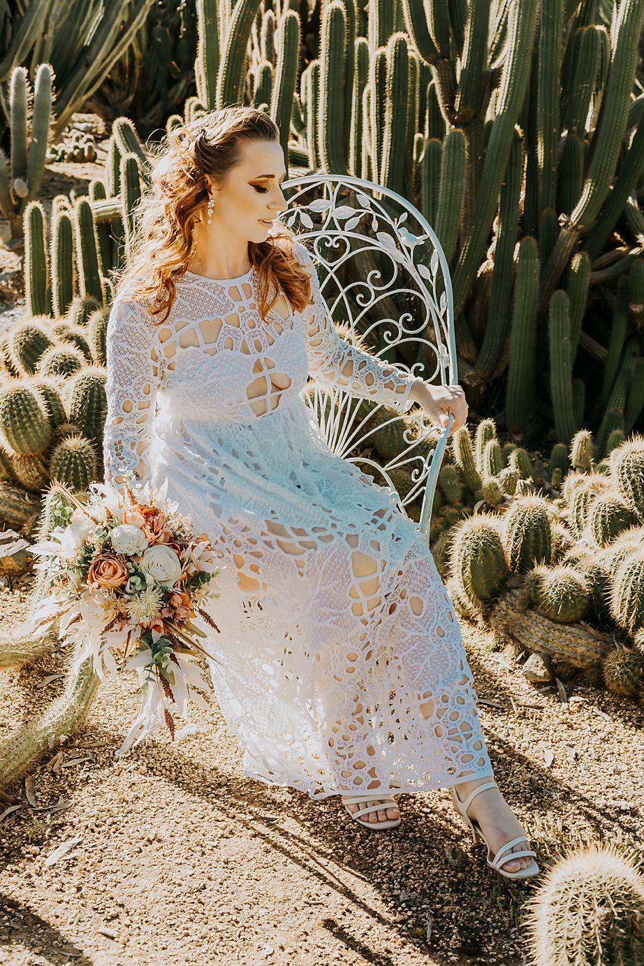 A delicate lace wedding dress glows in sunlight among desert cacti as a figure sits in an ornate white chair.