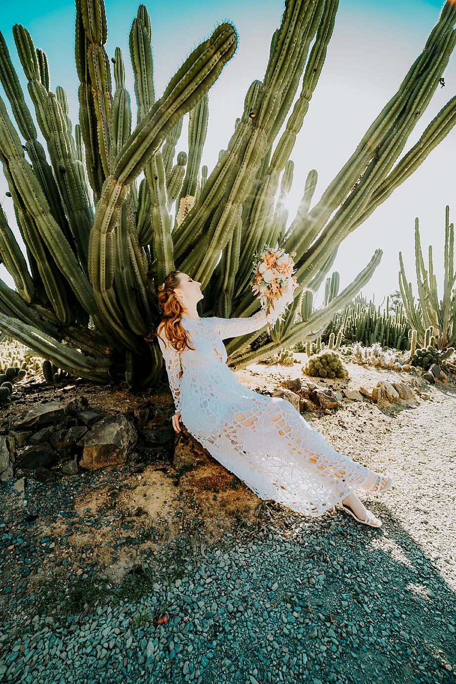 A flowing white dress cascades near tall desert cactus plants against a bright turquoise sky in a dreamy desert scene.