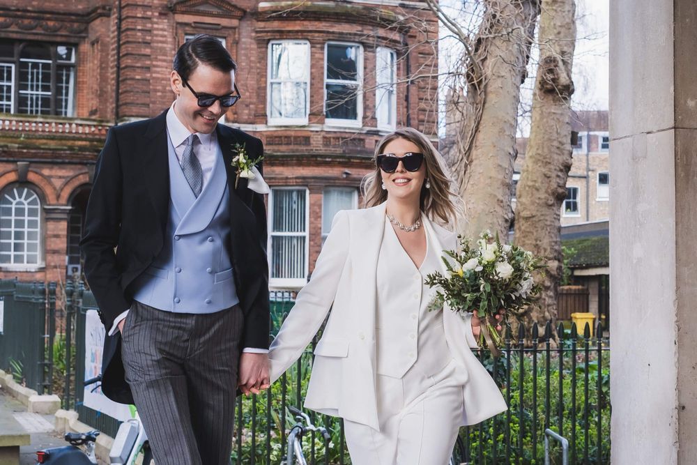 Stylish couple in formal attire walking together on city sidewalk with bride holding white flower bouquet.