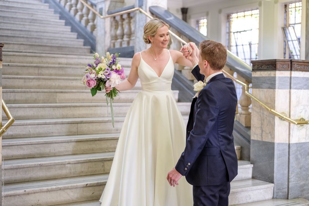 A bride and groom embrace on an elegant marble staircase with decorative flowers adorning the venue.