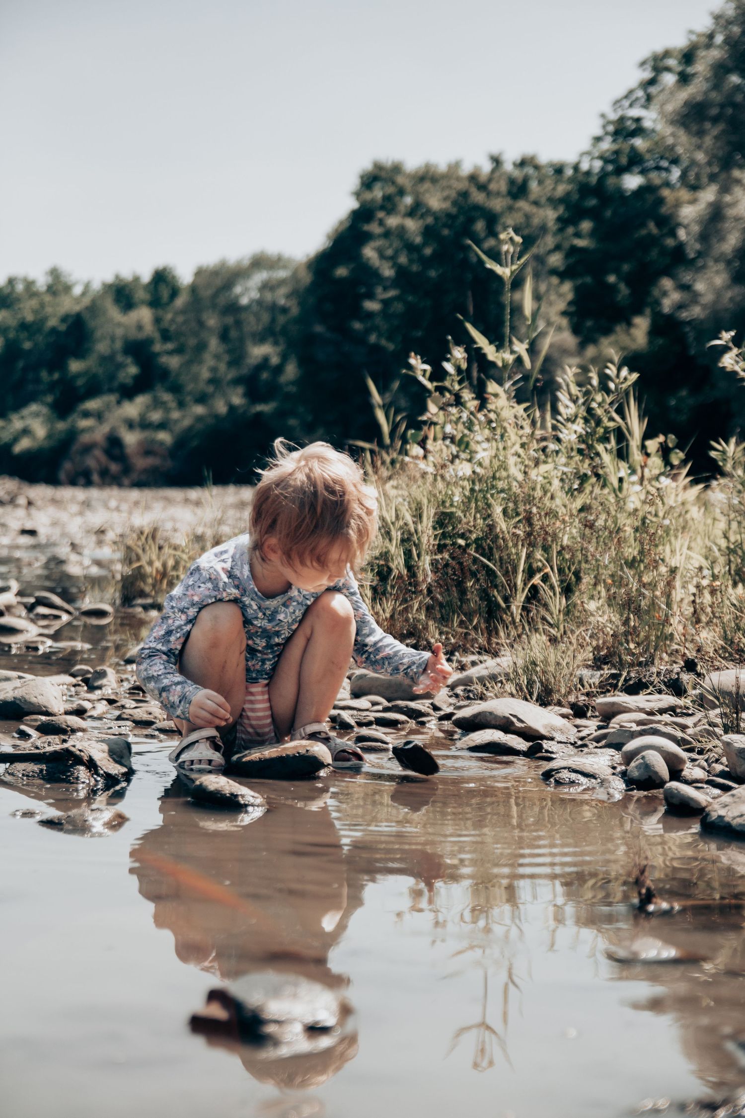 Child in patterned shirt leaning over to touch the water's surface near a rocky shore.