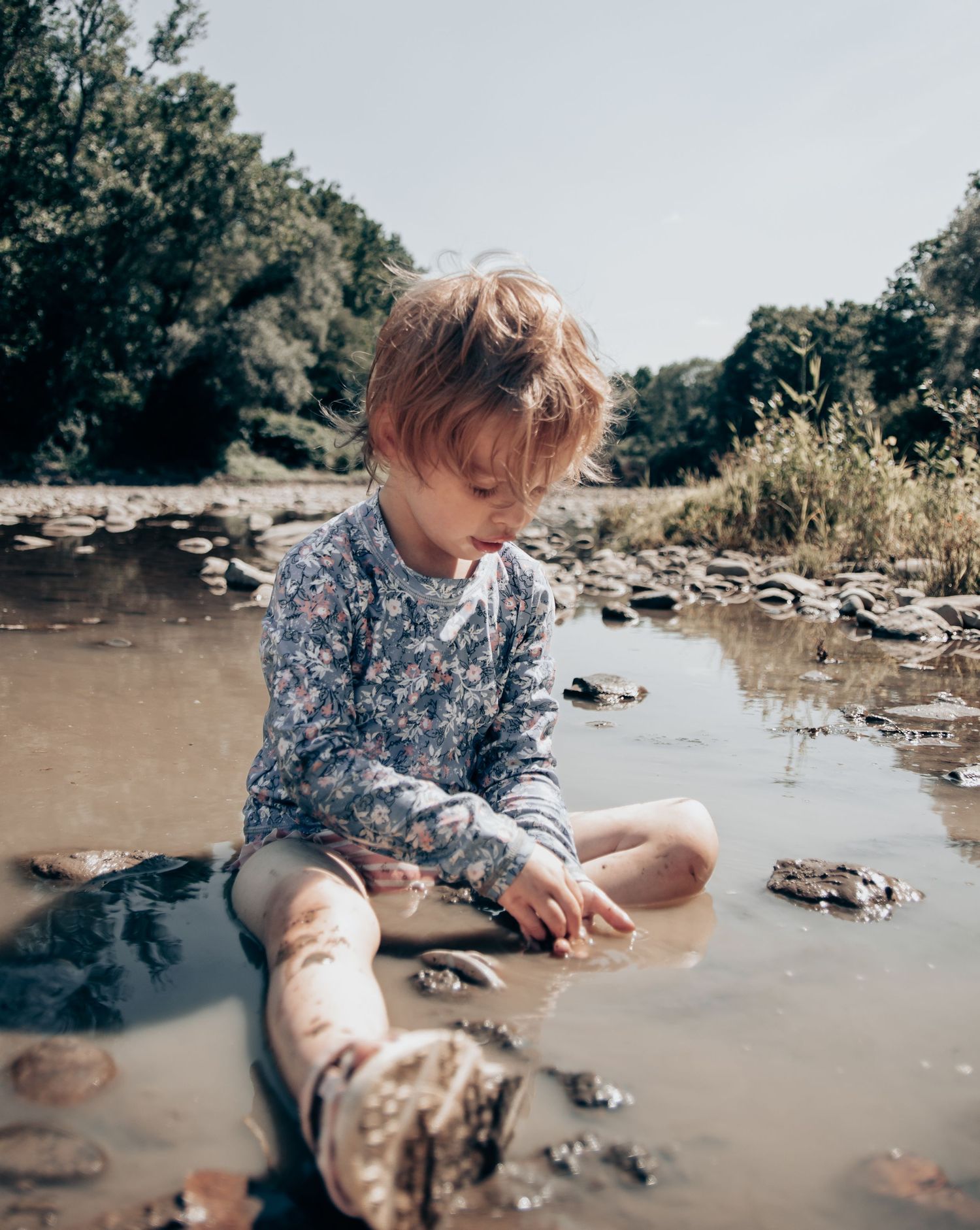 Child in floral shirt playing in shallow muddy water near rocks.