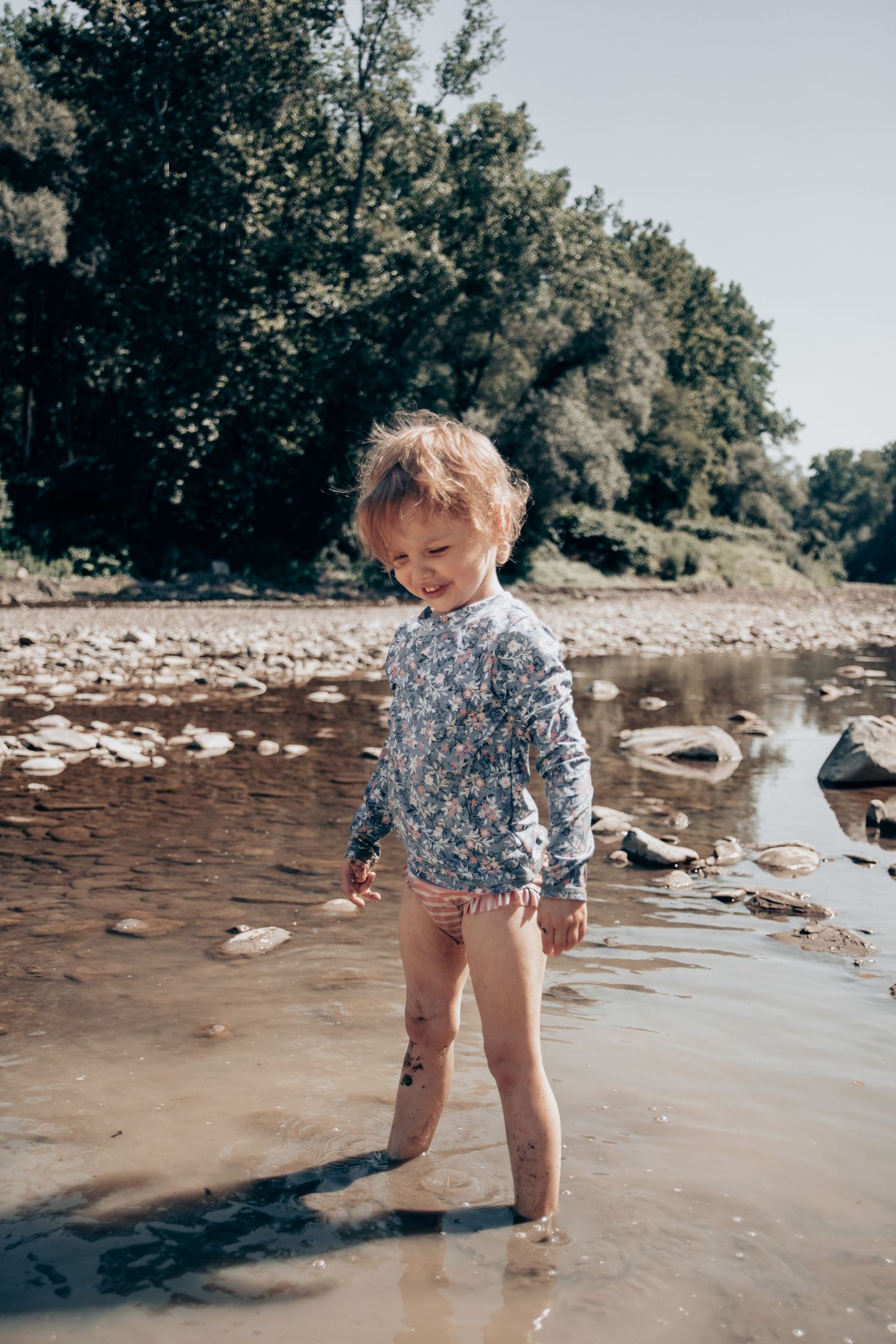Child in blue floral shirt walking through shallow water at rocky beach.