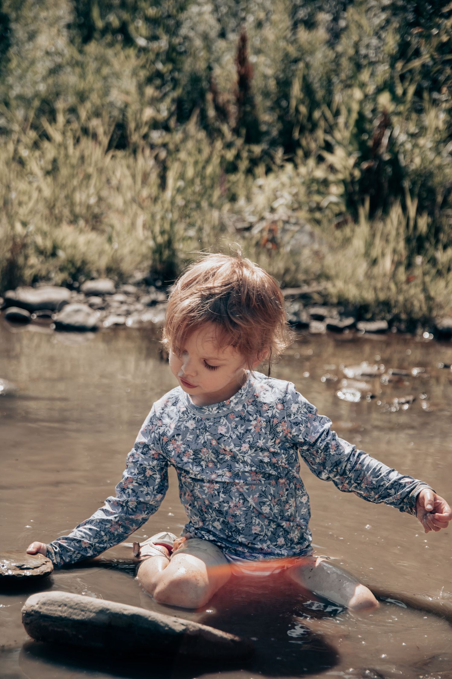 Child sitting and splashing in shallow muddy water wearing long sleeve floral shirt.