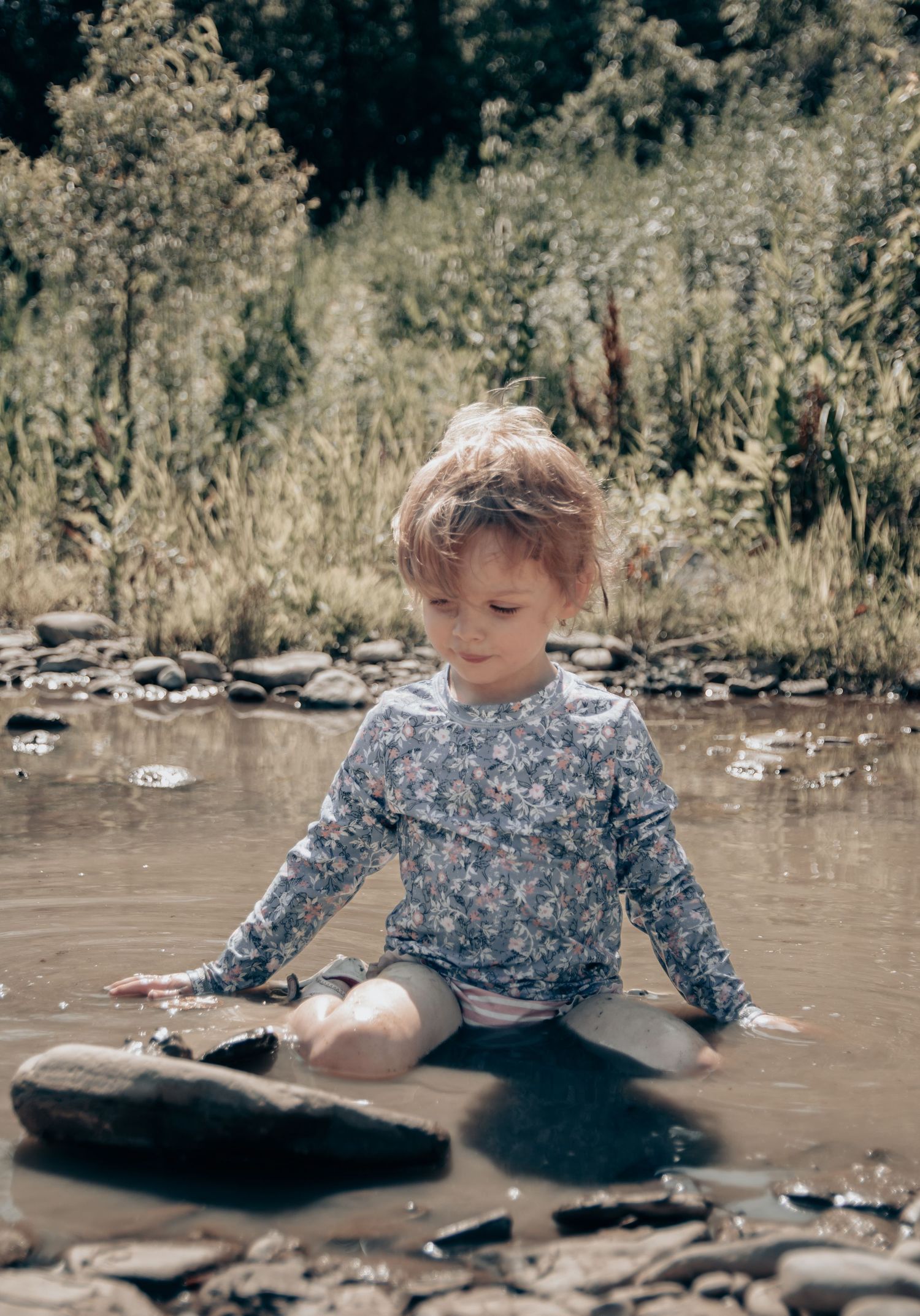 Child in floral shirt sitting and playing in shallow muddy water surrounded by rocks.