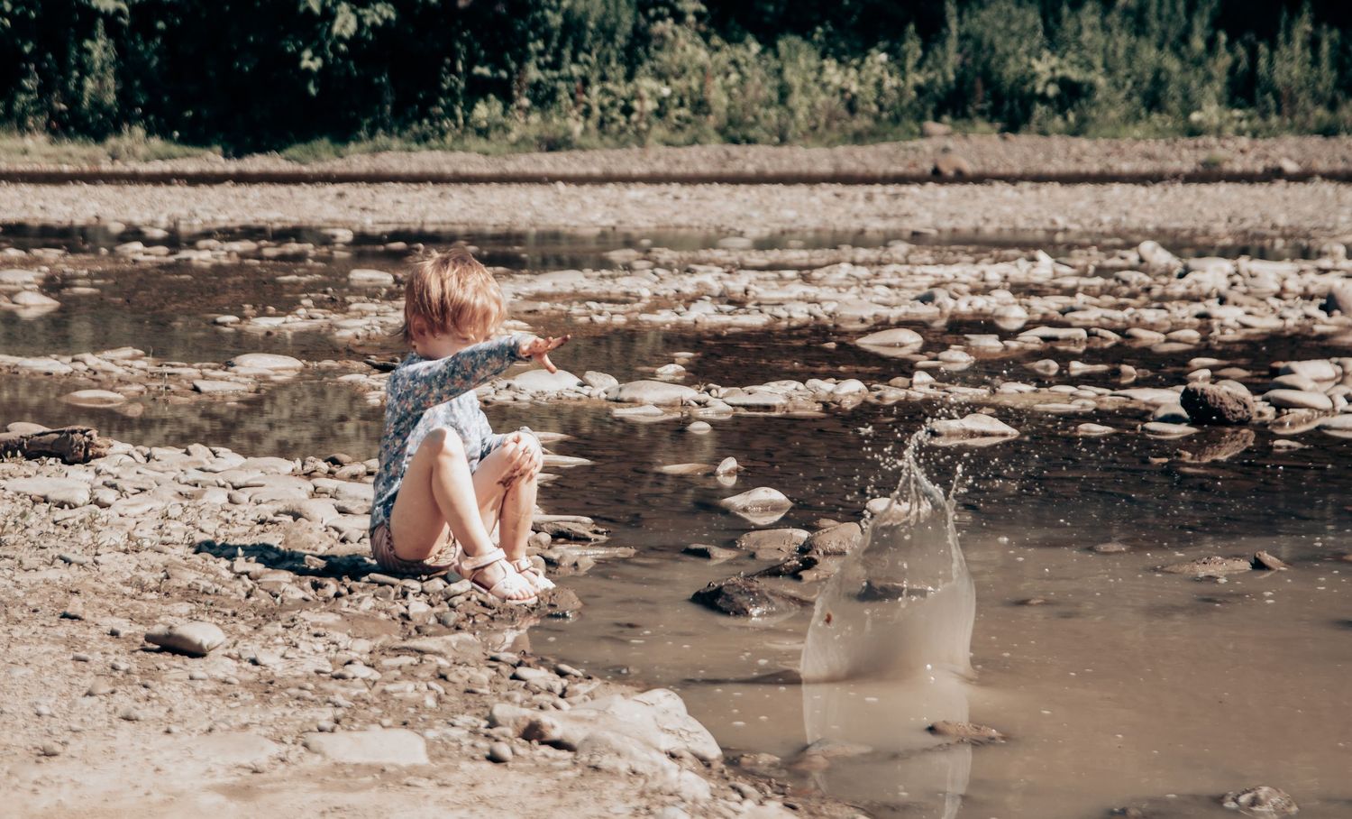 A person in casual attire sits by a rocky riverbed on a sunny day.