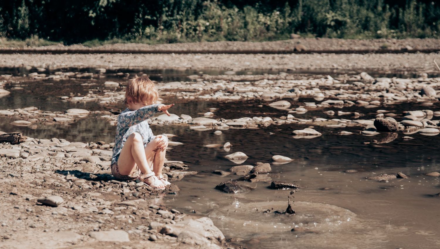 A person in casual attire sits by a rocky riverbed on a sunny day.