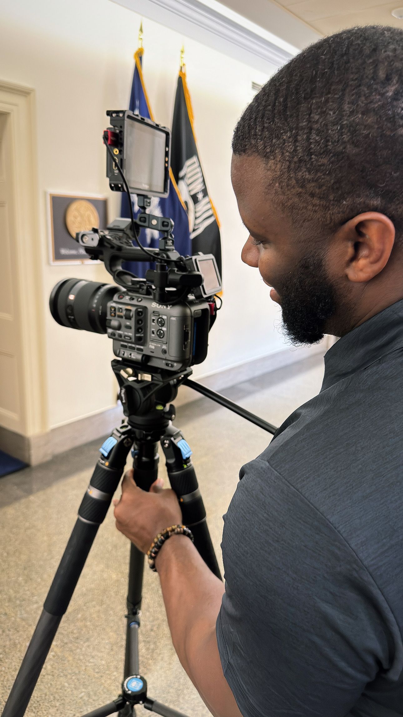 A person operates a professional video camera mounted on a tripod in a hallway setting.