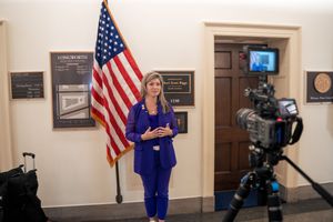 Woman in blue suit speaking in front of American flag while being filmed by professional camera in office hallway.