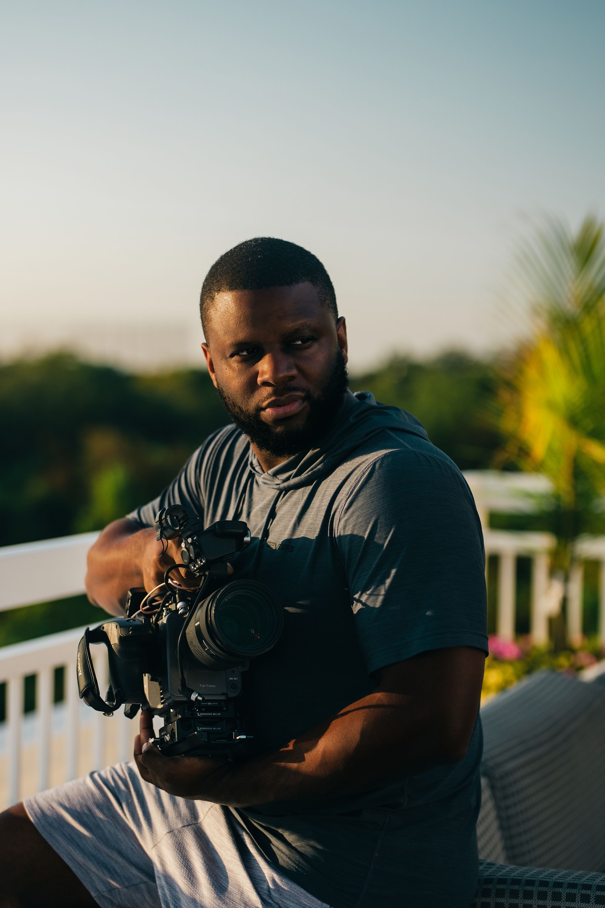 A videographer holds a professional camera while sitting on an outdoor deck at sunset.