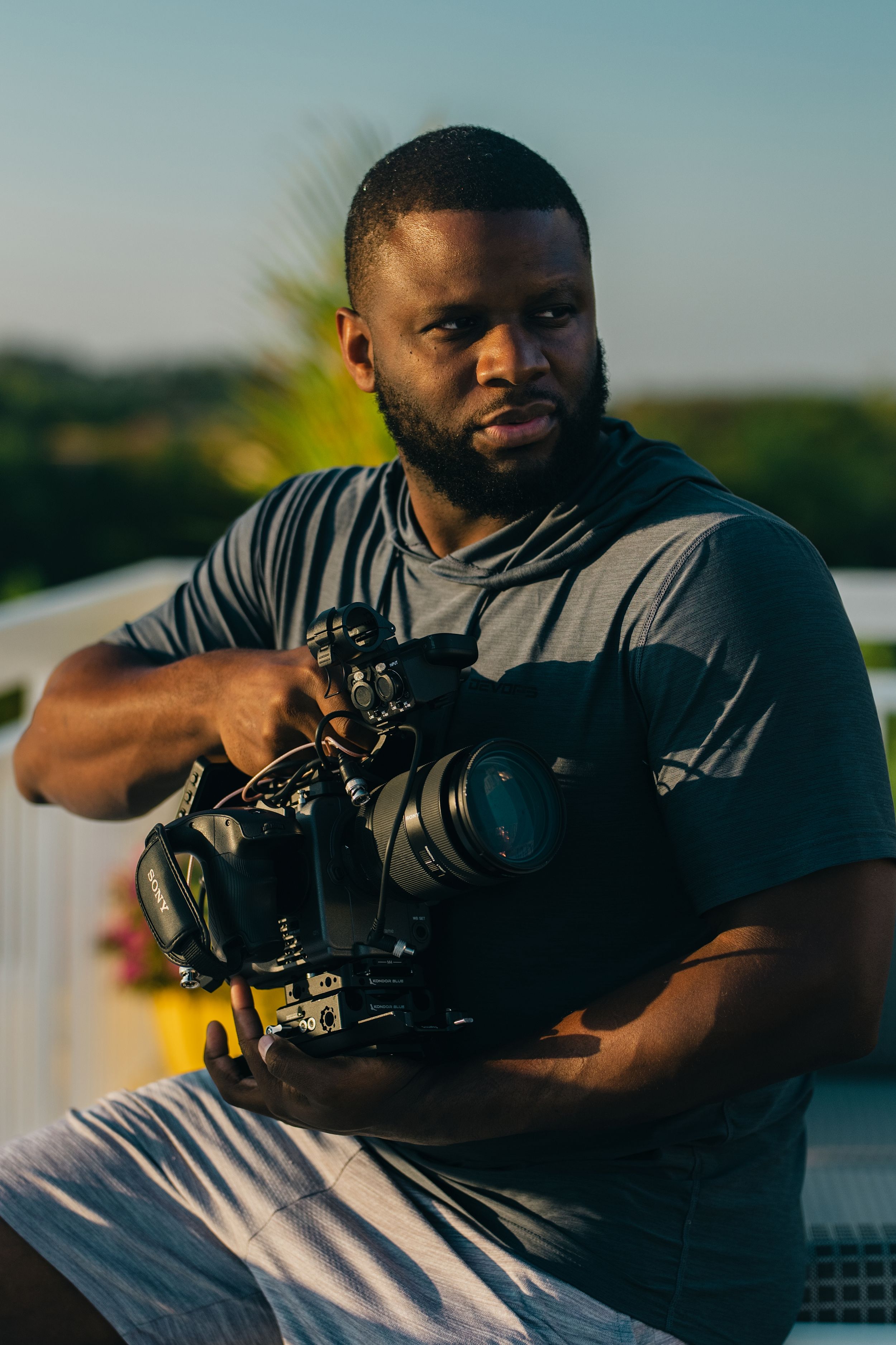 A photographer in a gray shirt holds a camera while taking photos at sunset by a lake.