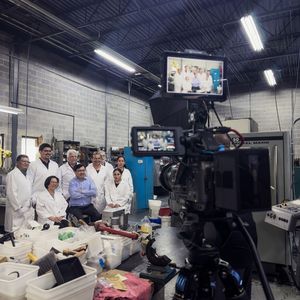 Professional video camera filming a group of people in white lab coats working in a laboratory setting.