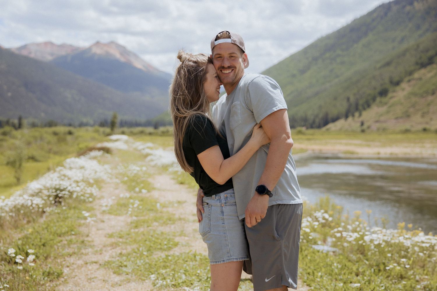 A couple embraces on a mountain path with scenic peaks and a winding river in the background.