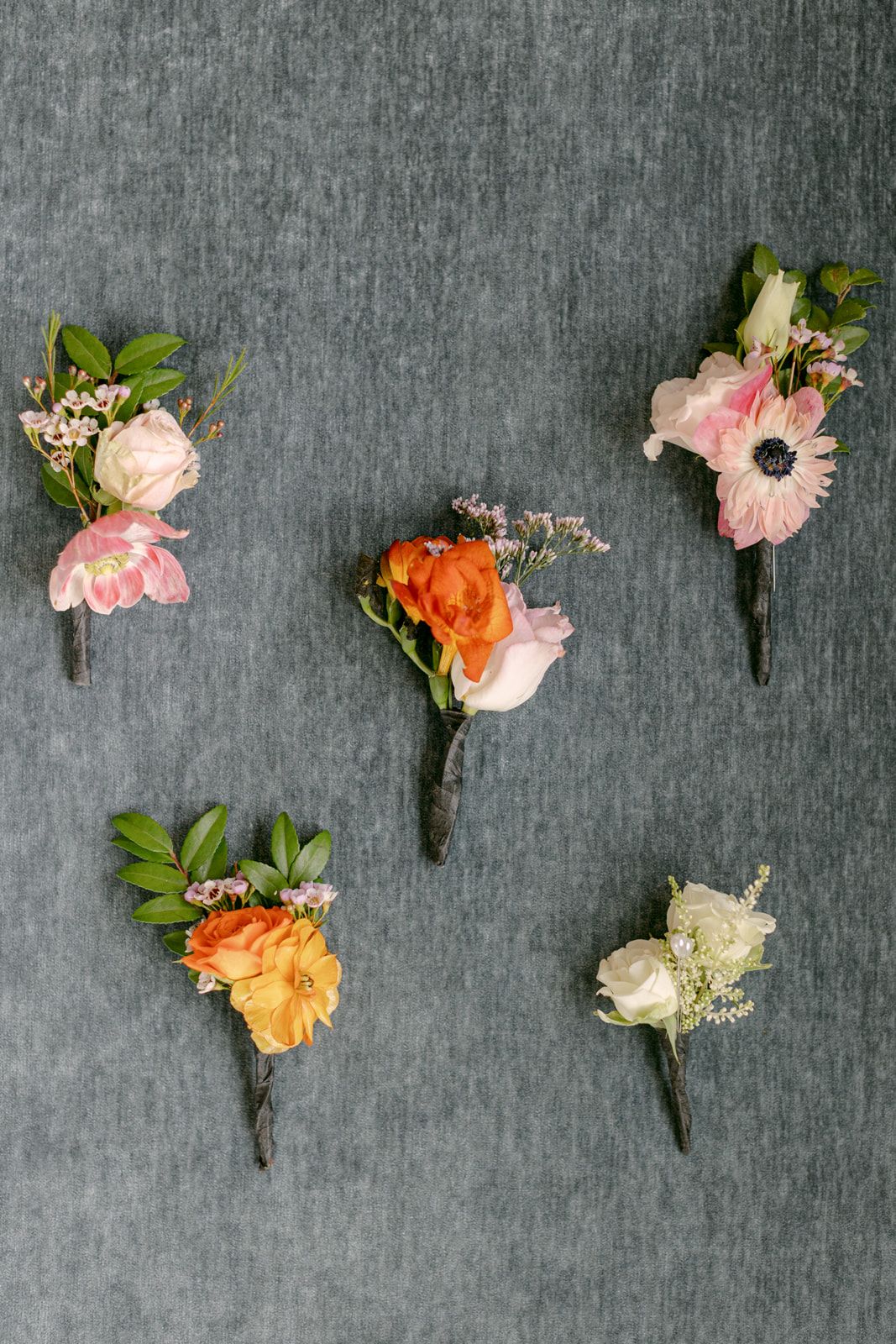 A collection of pink, peach and orange floral boutonnieres arranged in a line on a grey textured background.