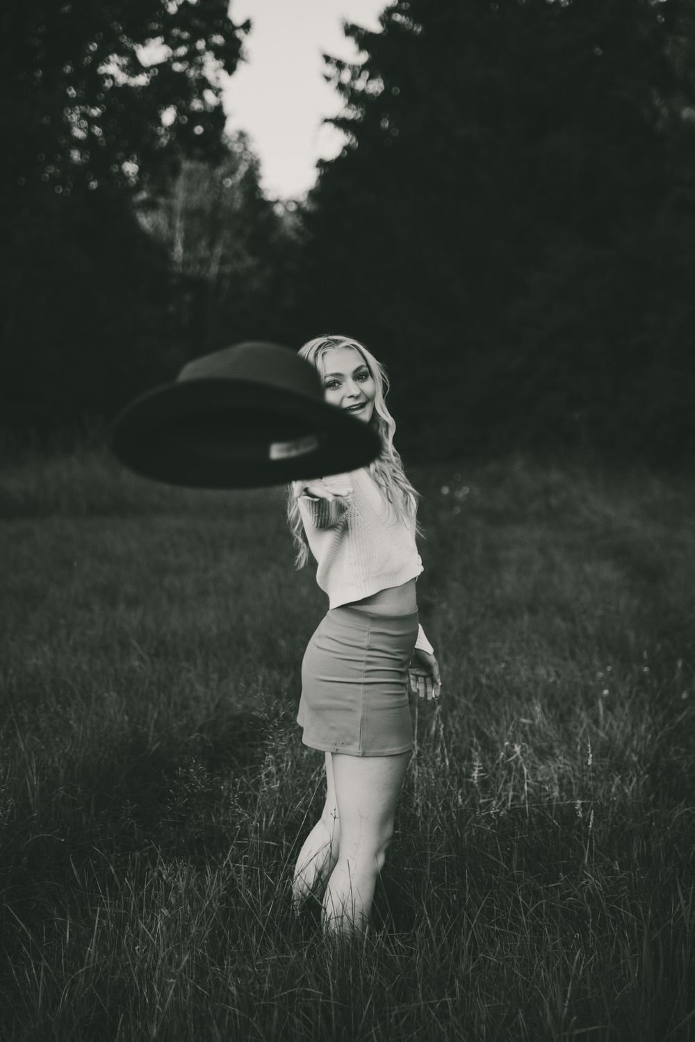 A black and white artistic portrait of someone playfully tossing a hat in a field.