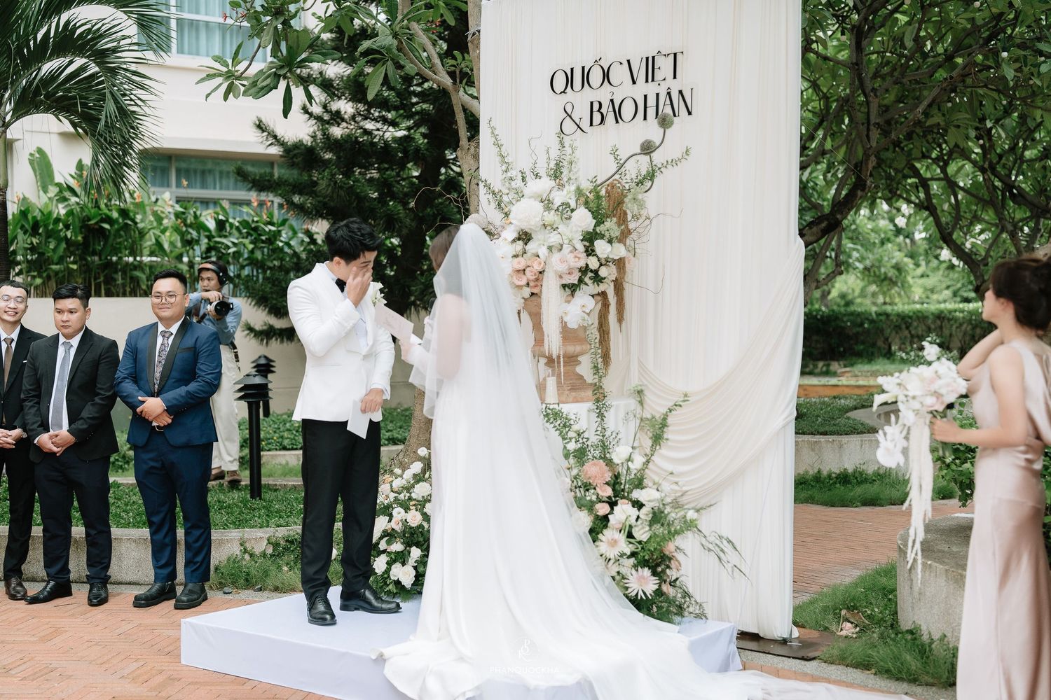 Groom leans in to kiss bride at outdoor wedding ceremony decorated with white floral arch backdrop.
