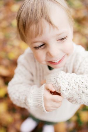 A child in a cozy cream sweater smiles while looking down at fallen autumn leaves.